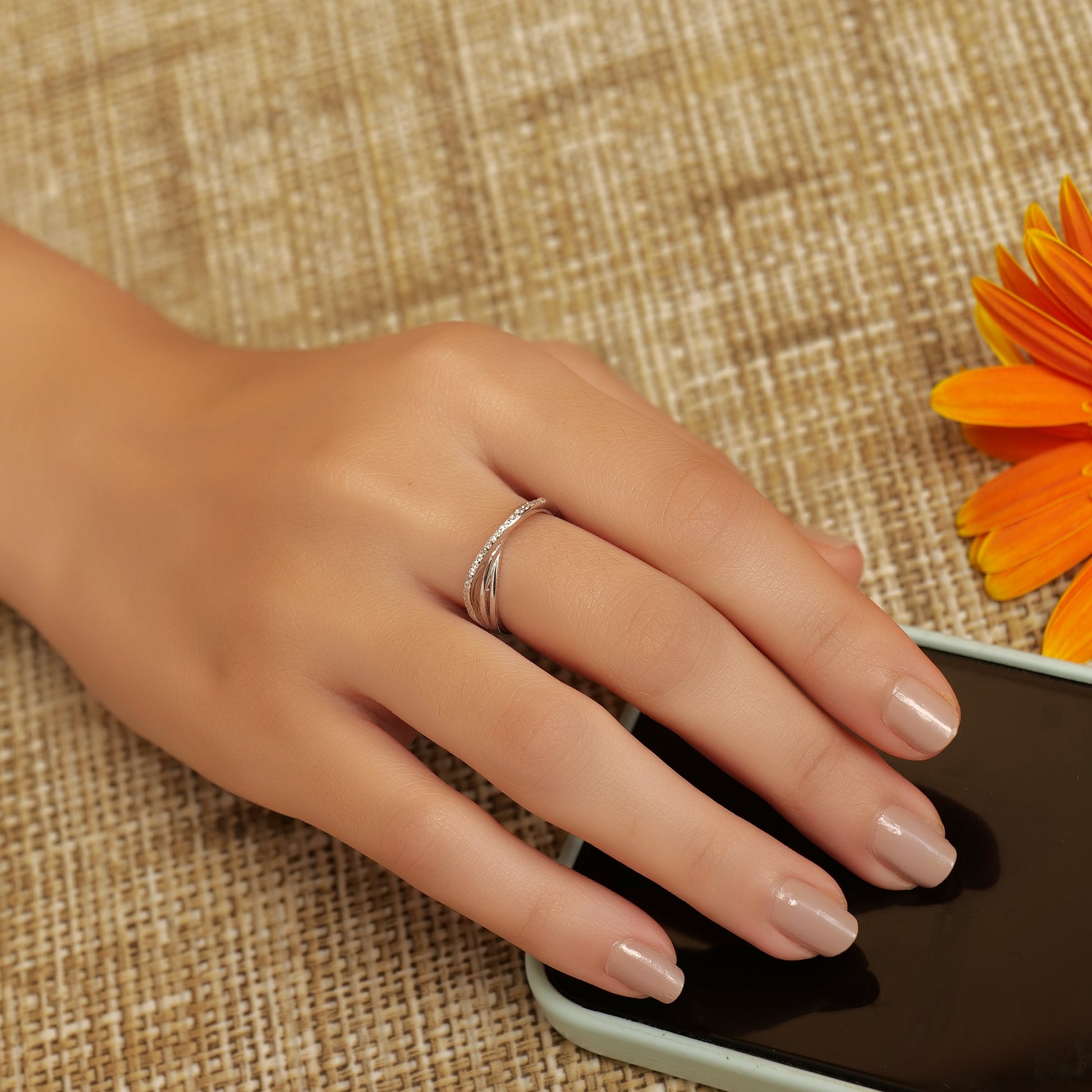 Hand with a silver ring on a smartphone against a textured brown background with an orange flower.
