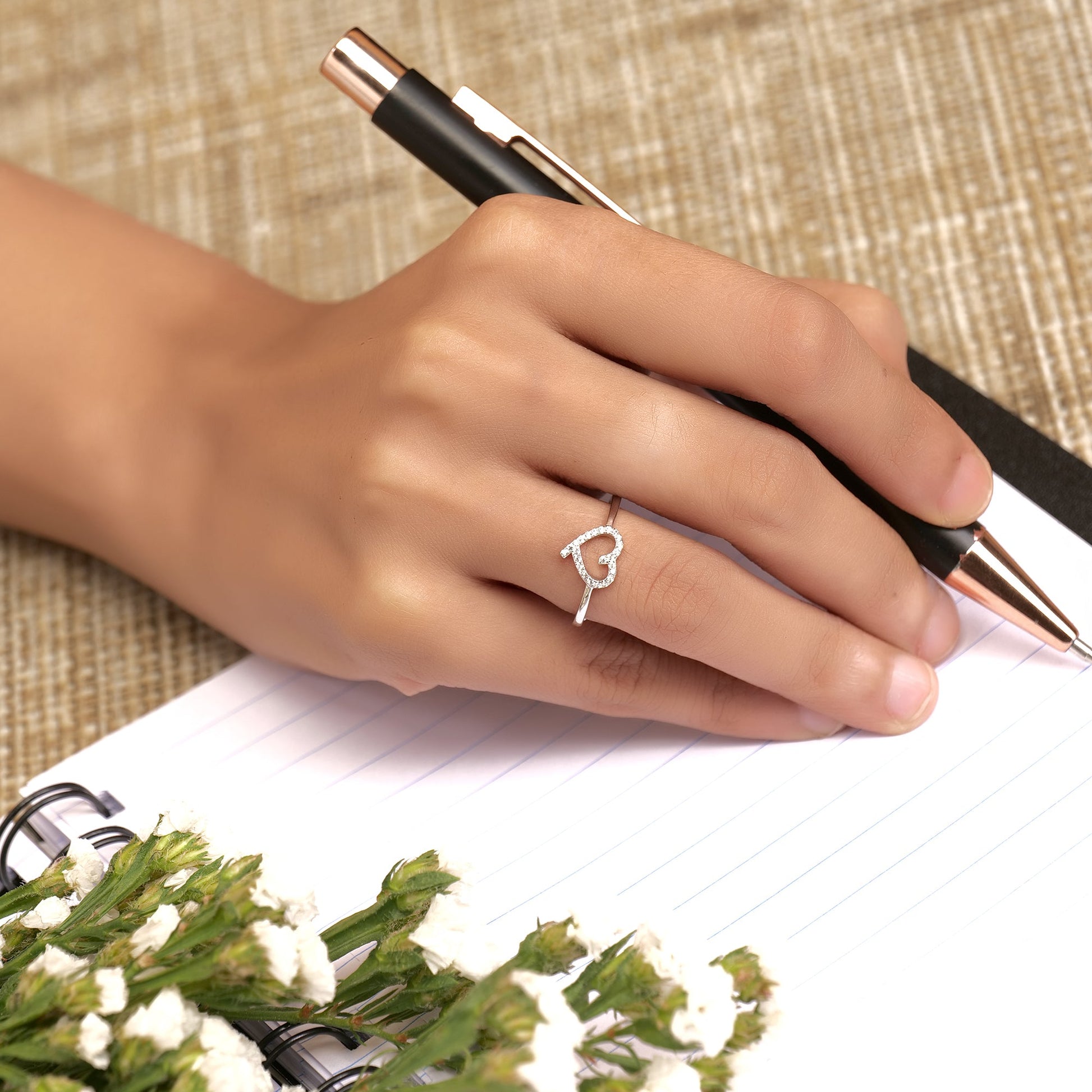 Hand holding a pen with a heart-shaped ring on a notebook with flowers